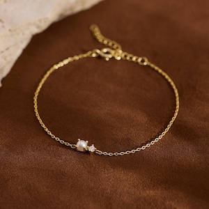 May include: A fine gold bracelet featuring a small square pearl and a tiny diamond. The bracelet is displayed on a brown surface. The Knot Knot Co. is written in the bottom right corner.