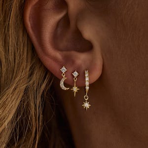 May include: Three gold earrings with dangling charms. The first earring features a crescent moon with a small crystal. The second earring features a star with a small crystal. The third earring features a larger star with a crystal and a hoop.