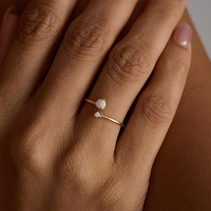 Könnte beinhalten: Ein goldfarbener Ring mit zwei weißen Opalsteinen. Der Ring hat ein zartes, offenes Design, mit einem Opal oben und einem kleineren darunter. The Knot Knot Co. steht unten rechts.