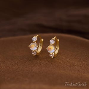 May include: A pair of gold-colored hoop earrings. Each earring features a central, iridescent, round stone, flanked by two smaller, clear, round stones. The earrings are set against a brown background.