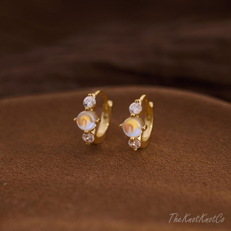 May include: A pair of gold-colored hoop earrings. Each earring features a central, iridescent, round stone, flanked by two smaller, clear, round stones. The earrings are set against a brown background.