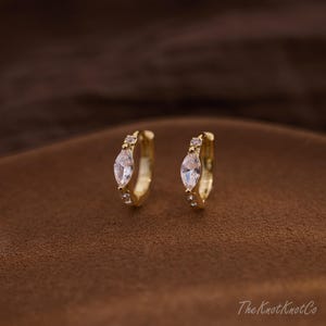 Può includere: Un paio di orecchini a cerchio color oro. Ogni orecchino presenta una pietra centrale trasparente a forma di marquise, affiancata da pietre trasparenti più piccole. Gli orecchini sono esposti su una superficie marrone.
