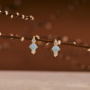 May include: A pair of gold-colored stud earrings. Each earring features a light blue, round gemstone in the center, flanked by two clear, round gemstones. The earrings are displayed against a brown background with a twig.