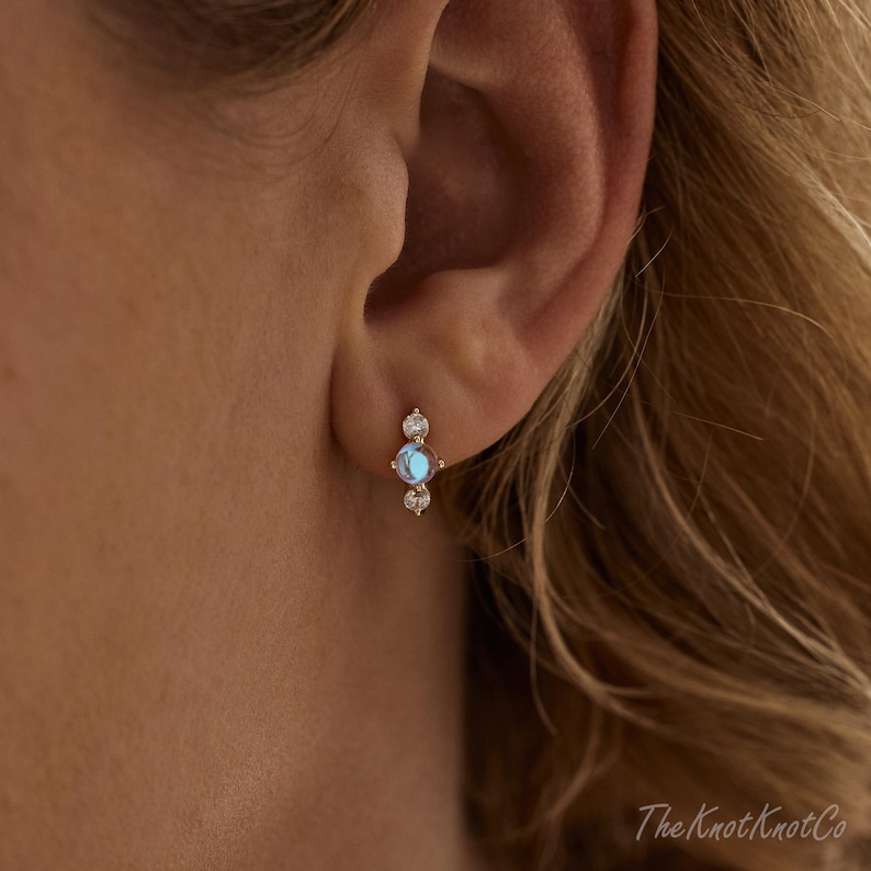 May include: A gold stud earring with a central blue opal stone and two smaller clear stones. The earring is worn on an ear, showcasing its delicate design and sparkle. The Knot Knot Co logo is in the bottom right corner.