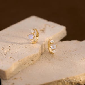 May include: A pair of gold-colored hoop earrings. Each earring features a central iridescent stone, flanked by two smaller, clear gemstones. The earrings are displayed on a textured, light-colored surface, with a blurred brown background.