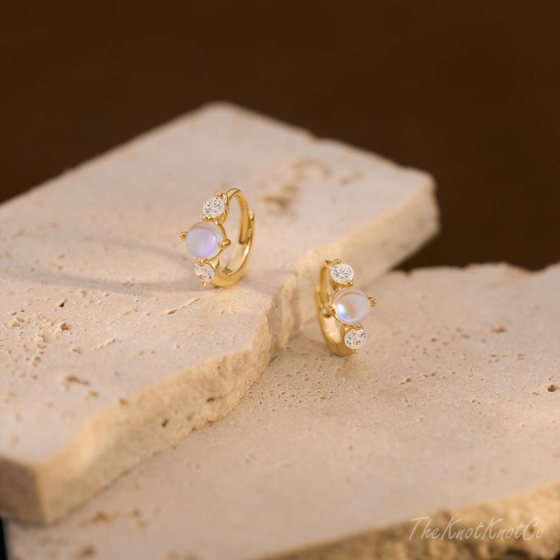 May include: A pair of gold-colored hoop earrings. Each earring features a central iridescent stone, flanked by two smaller, clear gemstones. The earrings are displayed on a textured, light-colored surface, with a blurred brown background.