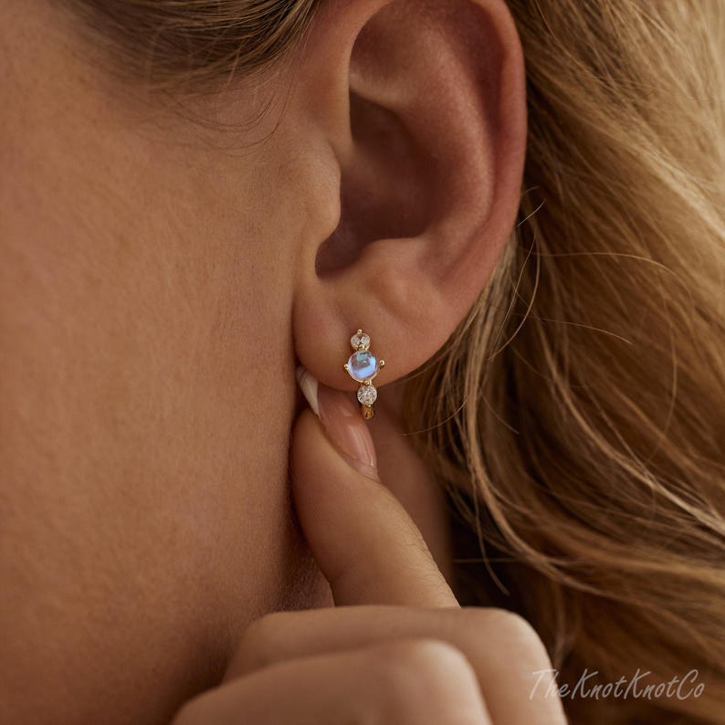 May include: A close-up of a gold earring with a small, round, light blue gemstone and two smaller clear gemstones. The earring is a hoop style and is being worn on an ear. The Knot Knot Co. is written in the bottom right corner.