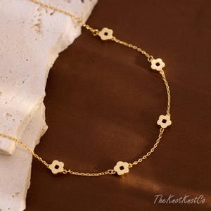 May include: A gold-coloured necklace with small, four-petal flower charms. The necklace is shown on a textured, off-white surface against a warm brown backdrop. The Knot Knot Co. is printed on the bottom right.
