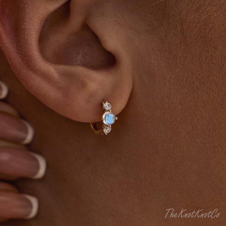 May include: A gold hoop earring with a central, round, light blue gemstone, flanked by two smaller, clear gemstones. The earring is set against the backdrop of a person's ear. The Knot Knotco logo is visible.