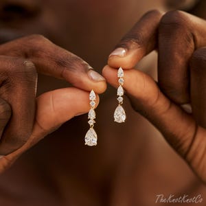 May include: A pair of gold drop earrings featuring a vertical arrangement of three clear, faceted stones. Each earring consists of a small round stone at the top, a marquise-shaped stone in the middle, and a large pear-shaped stone at the base.