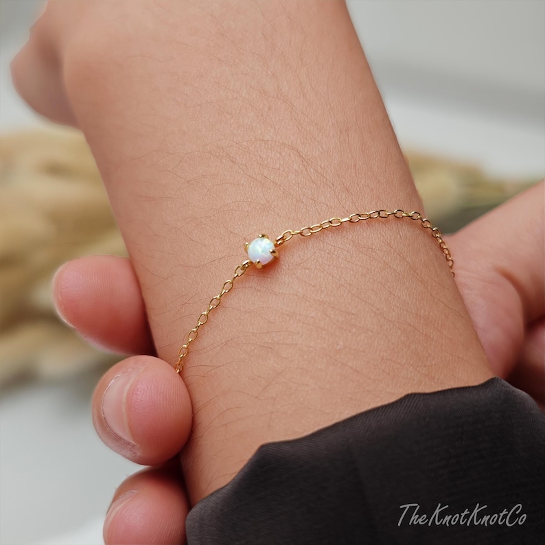 May include: A delicate gold chain bracelet with a single white opal gemstone. The bracelet is shown on a person's wrist.