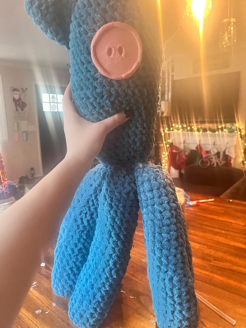 May include: A large, handmade blue crochet doll with a round, peach-colored button detail. The doll has three long, cylindrical legs and a textured, plush appearance. The doll is held up by a hand.