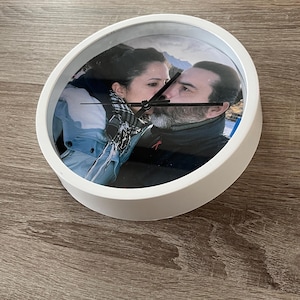 Può includere: Orologio rotondo bianco con una fotografia di una coppia che si bacia. L'orologio ha lancette nere e una cornice bianca. Lo sfondo è una superficie di legno.