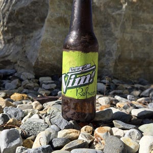 Peut inclure: Une bouteille en verre marron avec une étiquette verte et jaune indiquant "Vim! Refreshed", sur un fond de cailloux et de ciel bleu. La bouteille semble sale, comme si elle était à l'extérieur.