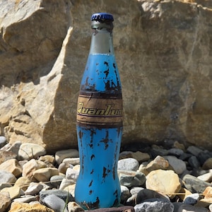 Op de afbeelding: Een blauwe glazen fles met een blauwe dop en een verweerd etiket met de tekst "Quantum". De fles staat tussen rotsen en kiezels, met een grote rotsformatie op de achtergrond. De lucht is blauw.