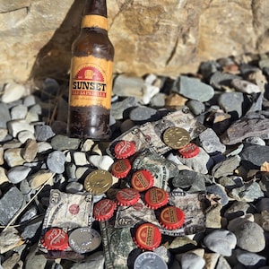 Puede incluir: Una botella de vidrio marrón de refresco Sunset Sarsaparilla con tapa roja, sobre un lecho de rocas, moneda, monedas y tapones. La botella tiene una etiqueta de papel con el nombre de la marca. La escena es al aire libre.