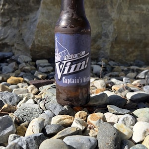May include: A brown glass bottle with a blue cap and a label that reads "You've got... Vim! Captain's blend." The bottle is on a bed of rocks with a large rock formation in the background under a blue sky.