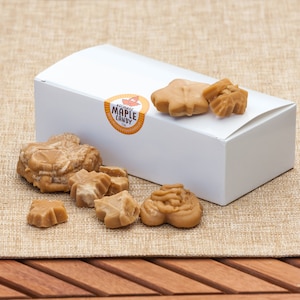 May include: A white box with a label that says "Authentic Maple Candy" and a variety of maple candies in the shape of leaves and a bear. The candies are on a brown burlap surface.