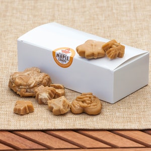 May include: A white box with a label that says "Authentic Maple Candy" and a variety of maple candies in the shape of leaves and hearts. The candies are on a brown burlap surface.
