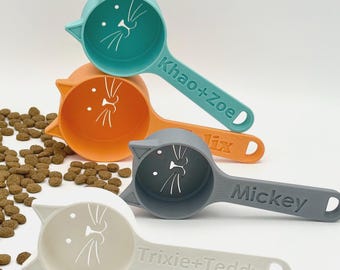 May include: Four cat-shaped pet food scoops in teal, orange, gray, and white. Each scoop features a cat face design inside the bowl and a handle personalized with names: Khao+Zoe, Felix, Mickey, and Trixie+Teddy. Small brown kibble pieces are scattered.