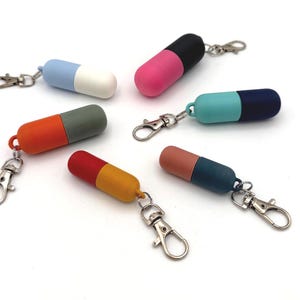 May include: Six pill-shaped keychains, each featuring a two-tone design with a metal lobster clasp. The colors include light blue and white, pink and black, teal and dark blue, orange and gray, red and yellow, and peach and dark blue.