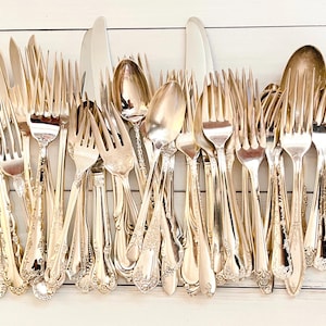 May include: A collection of vintage silver-plated flatware, including forks, knives, and spoons. The silverware has ornate, detailed handles and a shiny, reflective surface. The arrangement is on a white wooden surface.