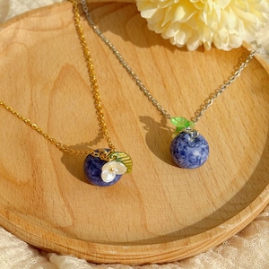 Könnte beinhalten: Zwei Halsketten mit Blaubeer-Anhängern. Eine hat eine goldene Kette und einen Anhänger mit einer weißen Blume und grünen Blättern. Die andere hat eine silberne Kette mit einem grünen Blatt und einem Blaubeer-Anhänger. Beide liegen auf einem Holztablett.