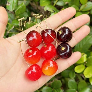 Könnte beinhalten: Ohrringe in Kirschform. Die Ohrringe zeigen rote und dunkelrote Harzkirschen mit goldfarbenen Stielen. Die Kirschen sind in einer Gruppe angeordnet und zeigen ihre glänzende Oberfläche und lebendigen Farben. Die Ohrringe sind ein einzigartiges Accessoire.