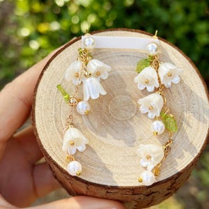 Op de afbeelding: Hangende oorbellen met witte bloemhangers, parelaccenten en goudkleurige kettingen. De oorbellen hebben kleine groene blad details, tegen een houten achtergrond.
