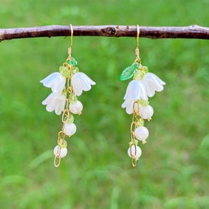 Könnte beinhalten: Zarte Blumenohrringe mit weißen Blumenanhängern, grünen Perlen und hängenden weißen Perlen. Die Ohrringe sind an goldfarbenen Haken und Ketten aufgehängt und schaffen ein anmutiges, von der Natur inspiriertes Design. Die Ohrringe hängen an einem braunen Ast.