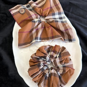 May include: Two brown plaid scrunchies and a matching fabric headband with a button, displayed on a white decorative plate. The scrunchies and headband feature a classic plaid pattern in shades of brown, white, and burgundy.