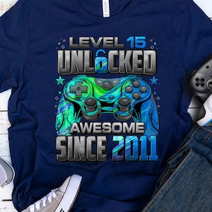 Pu&ograve; includere: T-shirt blu navy con grafica "Level 15 Unlocked Awesome Since 2011" e design controller blu e verde. Include un paio di scarpe da ginnastica grigie, due controller di gioco e un paio di jeans blu.