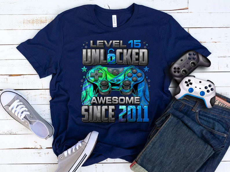 Pu&ograve; includere: T-shirt blu navy con grafica "Level 15 Unlocked Awesome Since 2011" e design controller blu e verde. Include un paio di scarpe da ginnastica grigie, due controller di gioco e un paio di jeans blu.