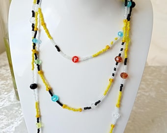 May include: A multi-strand beaded necklace displayed on a white mannequin. The necklace features yellow, black, white, and clear beads, with pops of color from red, green, and blue beads. One strand has a white bead with a red "F" on it.