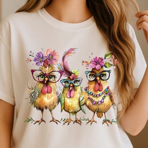 May include: Cream-colored t-shirt featuring a whimsical illustration of three chickens wearing glasses and floral headpieces. The chickens are in shades of yellow, orange, and green, with colorful accents. The design is printed on a soft, casual tee.