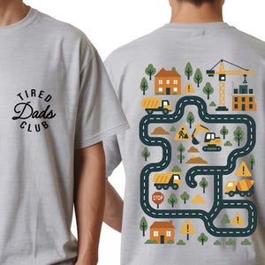 May include: Light gray t-shirt with the text "Tired Dads Club" in black script on the front. The back features a colorful construction-themed graphic with trucks, houses, trees, and a crane on a winding road.