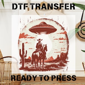 May include: A red, white, and blue DTF transfer design featuring a cowboy riding a horse in a desert landscape with a UFO hovering above. The text "READY TO PRESS" is printed below the design.