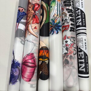 May include: A collection of cylindrical objects with various designs. One features an American flag, another a woman with a red bow, and others showcase tropical scenes, a pig, and text that reads "KLEIN". The objects have a white base and are likely decorative or functional.