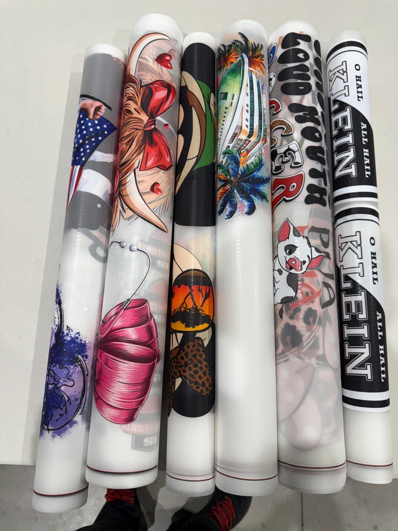 May include: A collection of cylindrical objects with various designs. One features an American flag, another a woman with a red bow, and others showcase tropical scenes, a pig, and text that reads "KLEIN". The objects have a white base and are likely decorative or functional.