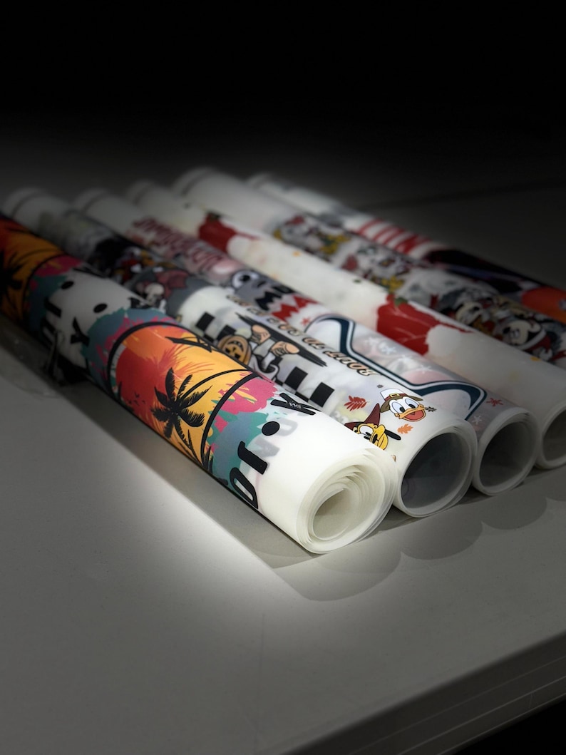 May include: Rolled-up colorful printed fabric rolls. The designs include tropical scenes, cartoon characters, and various patterns. The rolls are white with vibrant illustrations in shades of orange, blue, and green. The rolls are arranged in a row.
