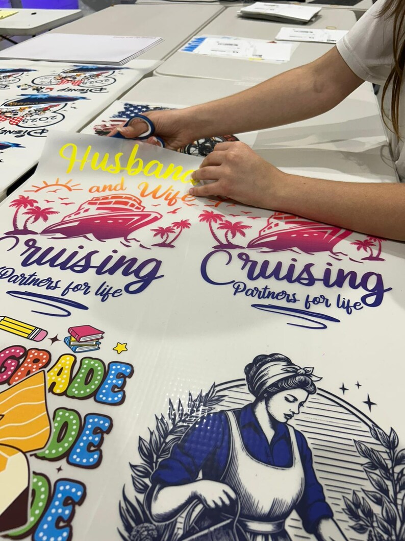 May include: A person is cutting a yellow vinyl decal that says "Husband and Wife" above a cruise ship graphic. The decal is for a shirt. Other designs include a school-themed graphic and a vintage-style woman.