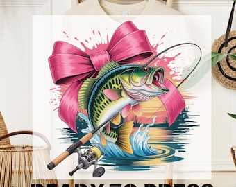 Puede incluir: Texto DTF TRANSFER y READY TO PRESS en una camiseta blanca. El diseño presenta una colorida ilustración de un róbalo saltando del agua, un lazo rosa y una caña de pescar. El pez tiene detalles verdes, amarillos y rosas.