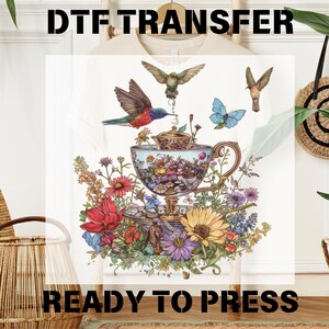 May include: A white t-shirt with a colorful design featuring a teacup filled with flowers and hummingbirds. The text "DTF TRANSFER READY TO PRESS" is printed on the bottom of the shirt.