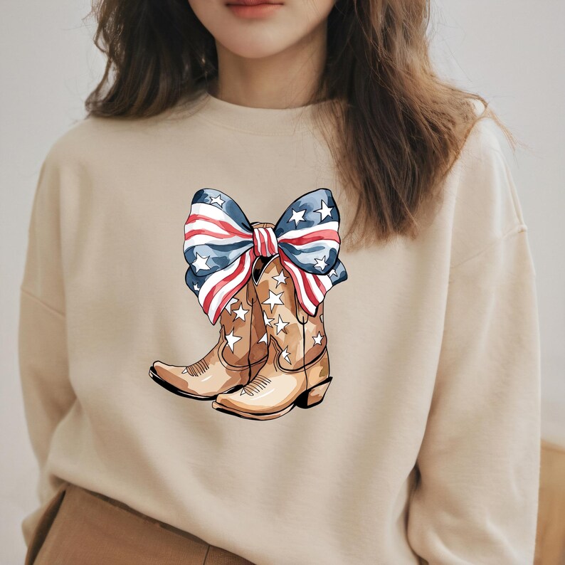 May include: A beige sweatshirt featuring a graphic of cowboy boots and a bow, both decorated with patriotic red, white, and blue colors. The boots have star accents. The bow is designed with stripes and stars.