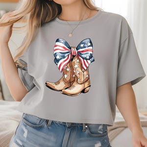 May include: A light gray t-shirt featuring a watercolor illustration of brown cowboy boots adorned with white stars. A large bow, colored with red and white stripes and blue stars, sits atop the boots. The shirt is a casual style.
