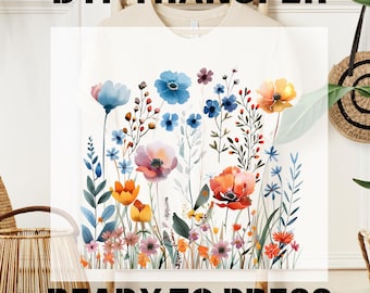 Puede incluir: Una camiseta color crema con un estampado floral estilo acuarela con flores silvestres azules, naranjas, rosas y rojas. El texto DTF TRANSFER READY TO PRESS aparece en letras negras en negrita en la parte superior e inferior.