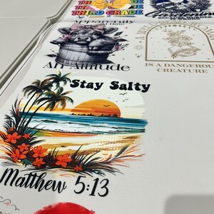May include: A collection of white printed sheets featuring various designs. Designs include text like "Stay Salty" and "Matthew 5:13", along with images of a cow, a sunset, and other graphics. The sheets are laid out on a white surface.
