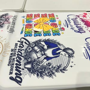 May include: White surface with various graphic designs. One design features a woman gardening with the text "Gardening because murder is wrong." Another design says "Third Grade" with a bow and pencils.