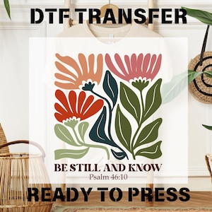 May include: A white t-shirt with a design of colorful flowers and the text "Be Still and Know, Psalm 46:10". The text "Ready to Press" is also visible below the design.