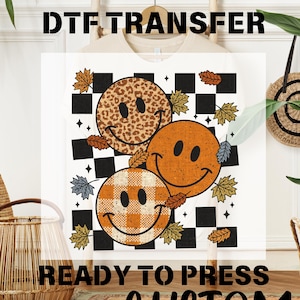 May include: White t-shirt with a DTF transfer design featuring three smiley faces with fall-themed patterns: leopard print, orange, and plaid. The design includes autumn leaves and a black and white checkered background. The text "READY TO PRESS" is visible.